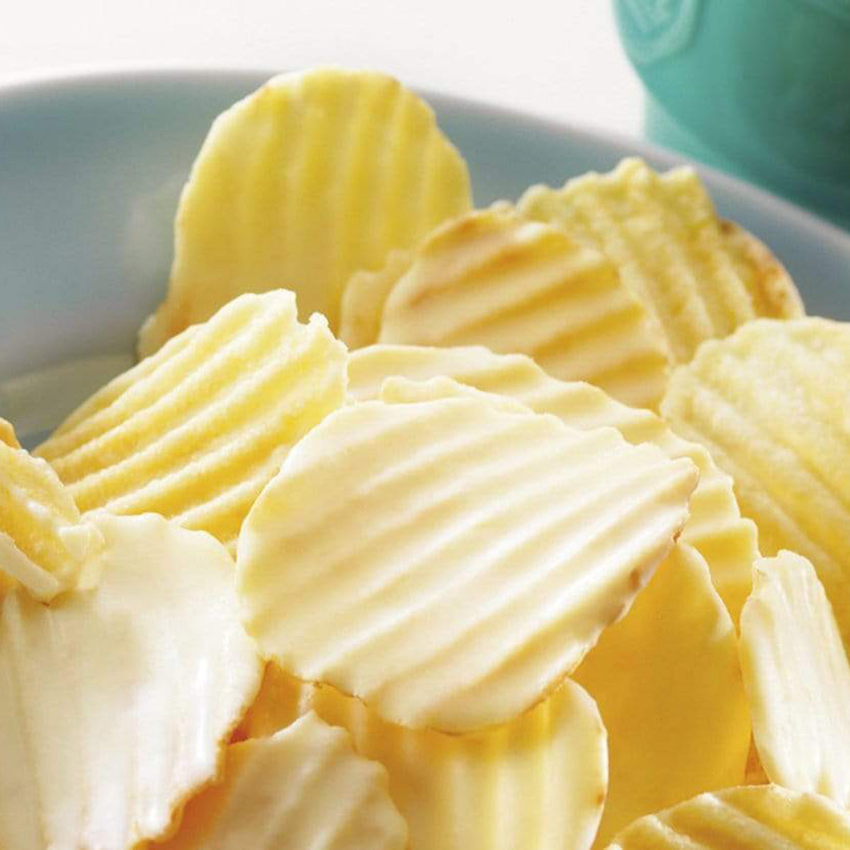 ROYCE' Chocolate - Potatochip Chocolate "Fromage Blanc" - Image shows white chocolate-coated potato chips with a ridged texture in a blue ceramic bowl. Background is white in color, and also seen is a portion of a ceramic cup on the right.