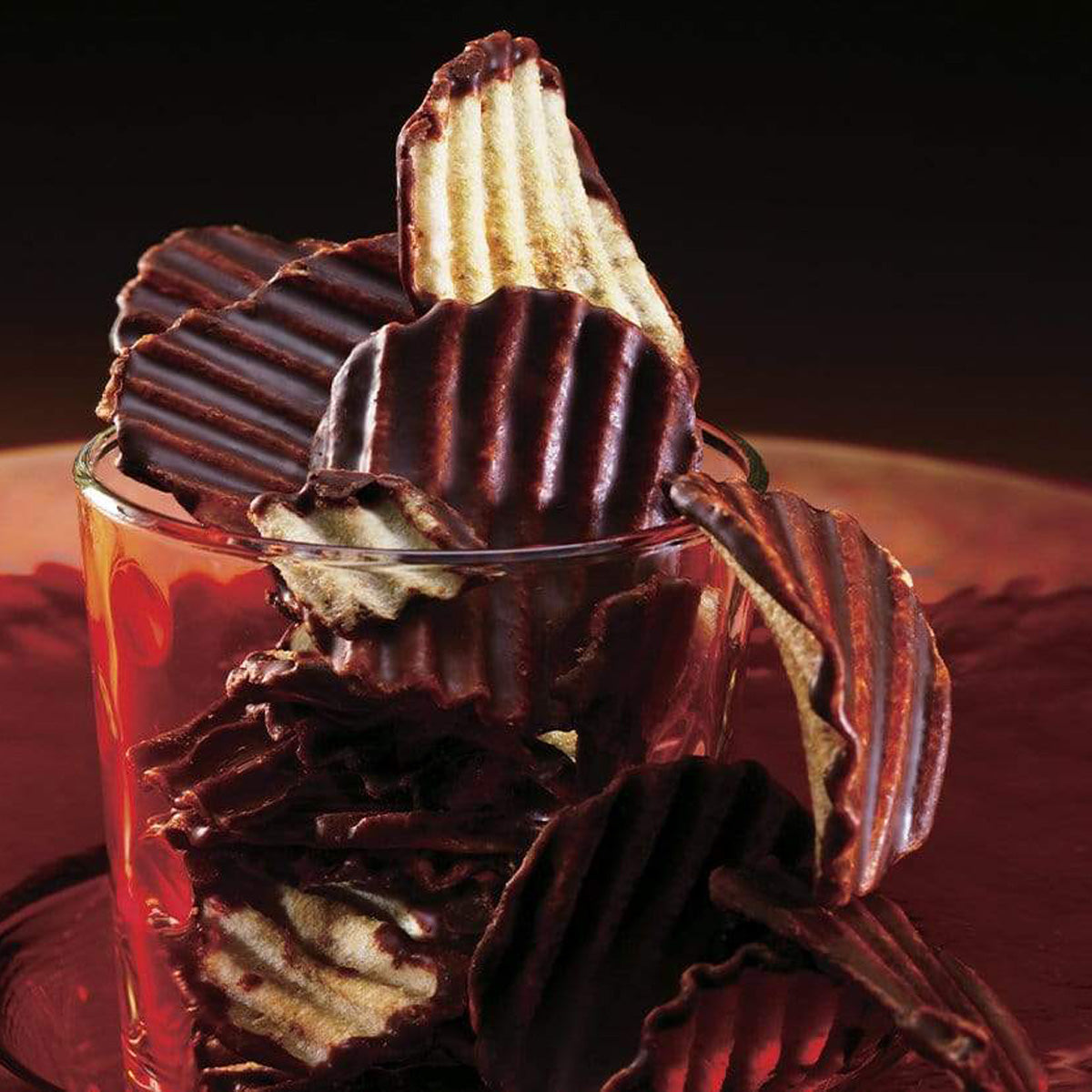 ROYCE' Chocolate - Potatochip Chocolate "Mild Bitter" - Image shows dark brown chocolate-covered potato chips with a ridged texture, as put inside a clear glass and on top of a brown plate. Background is in dark brown and black colors.