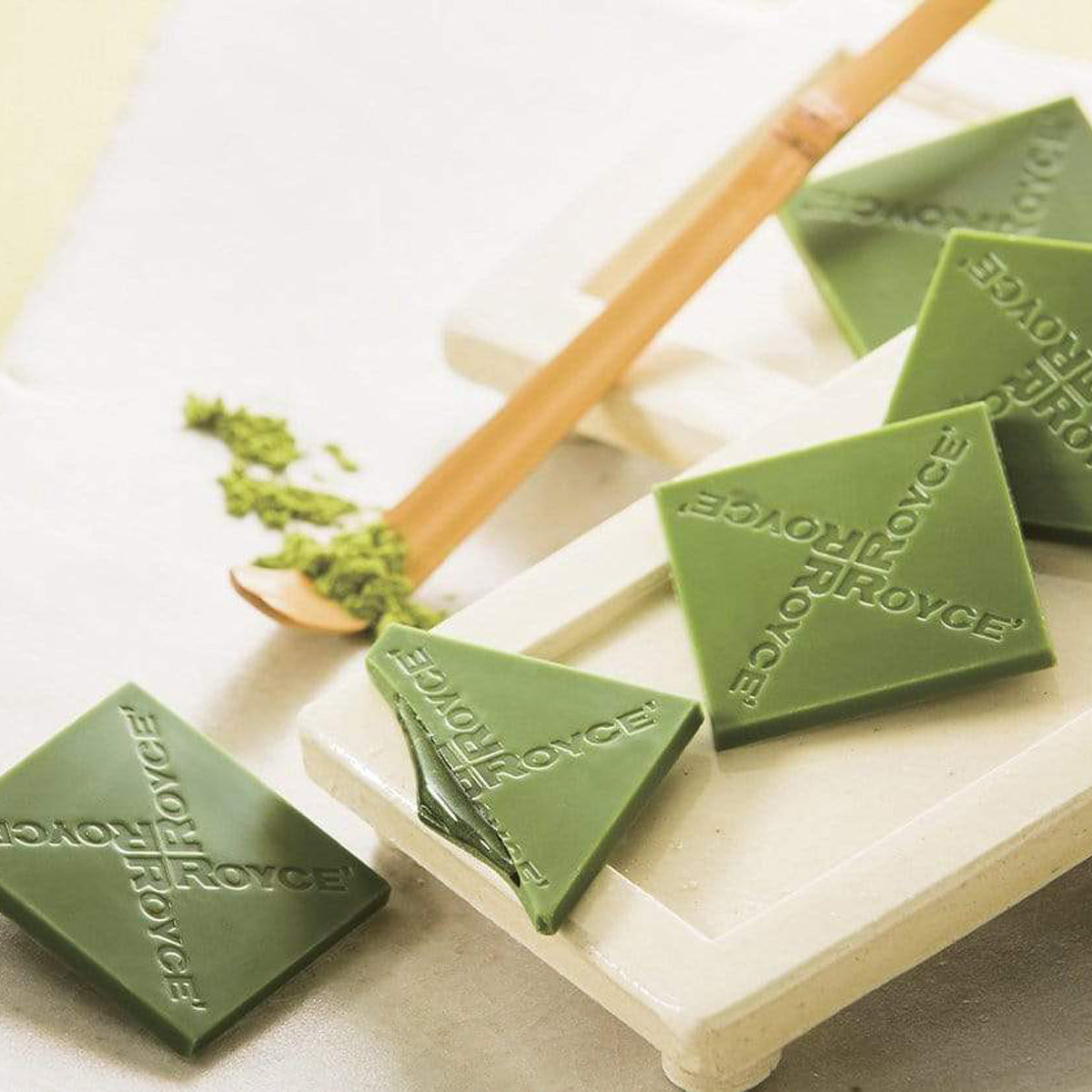 ROYCE' Chocolate - Prafeuille Chocolat "Matcha" - Image shows green chocolate squares filled with green sauce and engraved with the words "ROYCE'", as placed on white trays. Accents include a yellow wooden stick with green tea powder. Background is in white color.