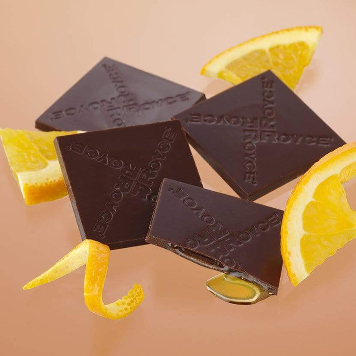 ROYCE' Chocolate - Prafeuille Chocolat "Orange" - Image shows brown chocolate squares filled with orange sauce and with the words "ROYCE'" engraved. Accents include fruit slices of orange and orange skin peel. Background is in color light brown.