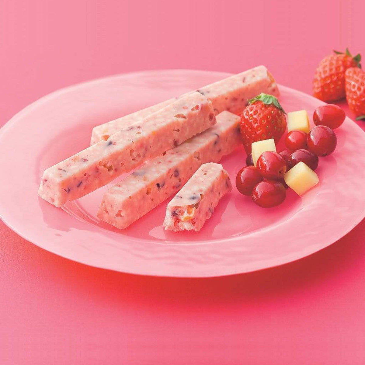 ROYCE' Chocolate - Fruit Bar Chocolate - Image shows pink chocolate bars on a pink plate with fruits such as red strawberries and grapes and yellow pineapple chunks. Background is in pink.