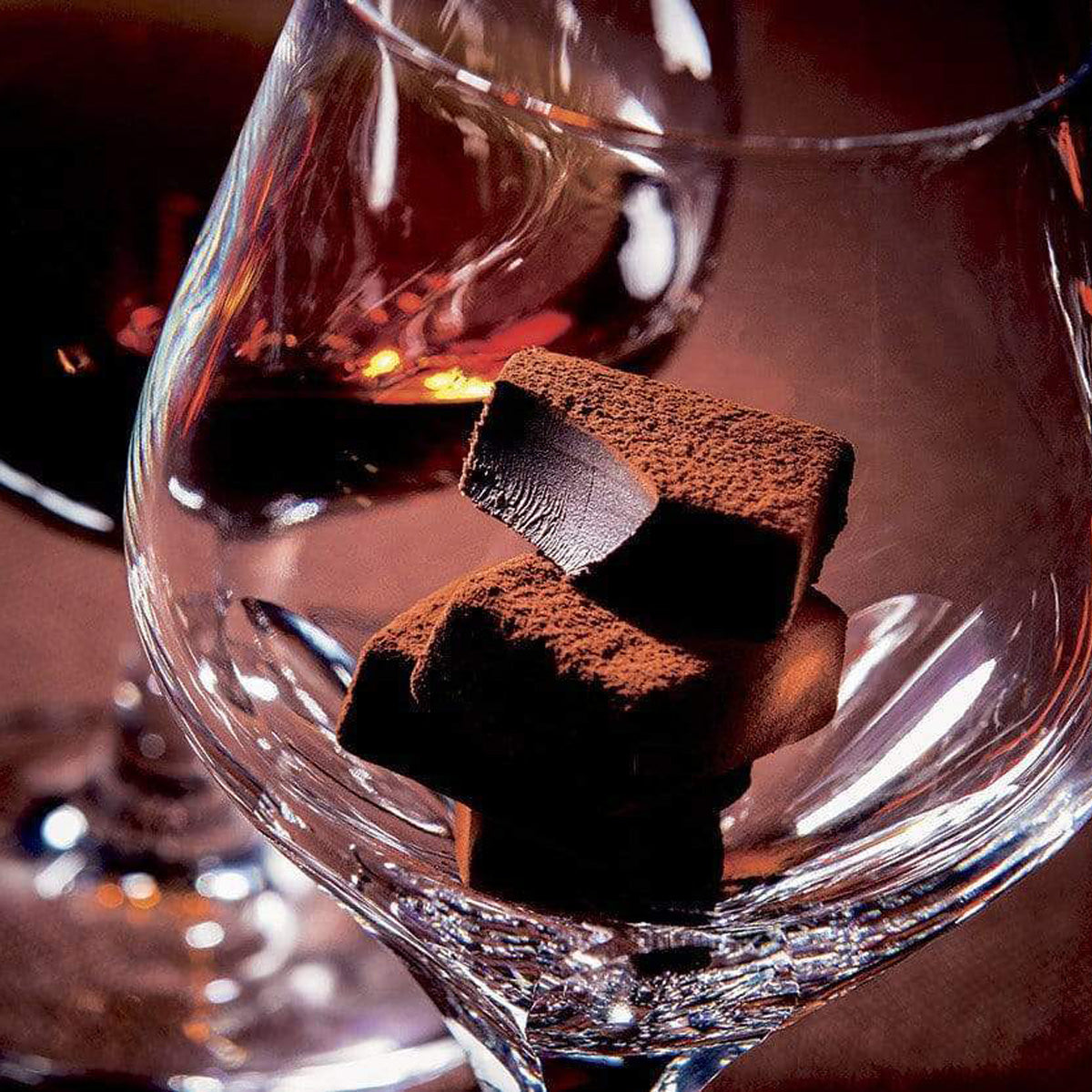 ROYCE' Chocolate - Nama Chocolate "Bitter" - Image shows brown blocks of chocolates inside a clear glass. Background is in dark brown and seen behind is another glass filled with amber-hued cognac.