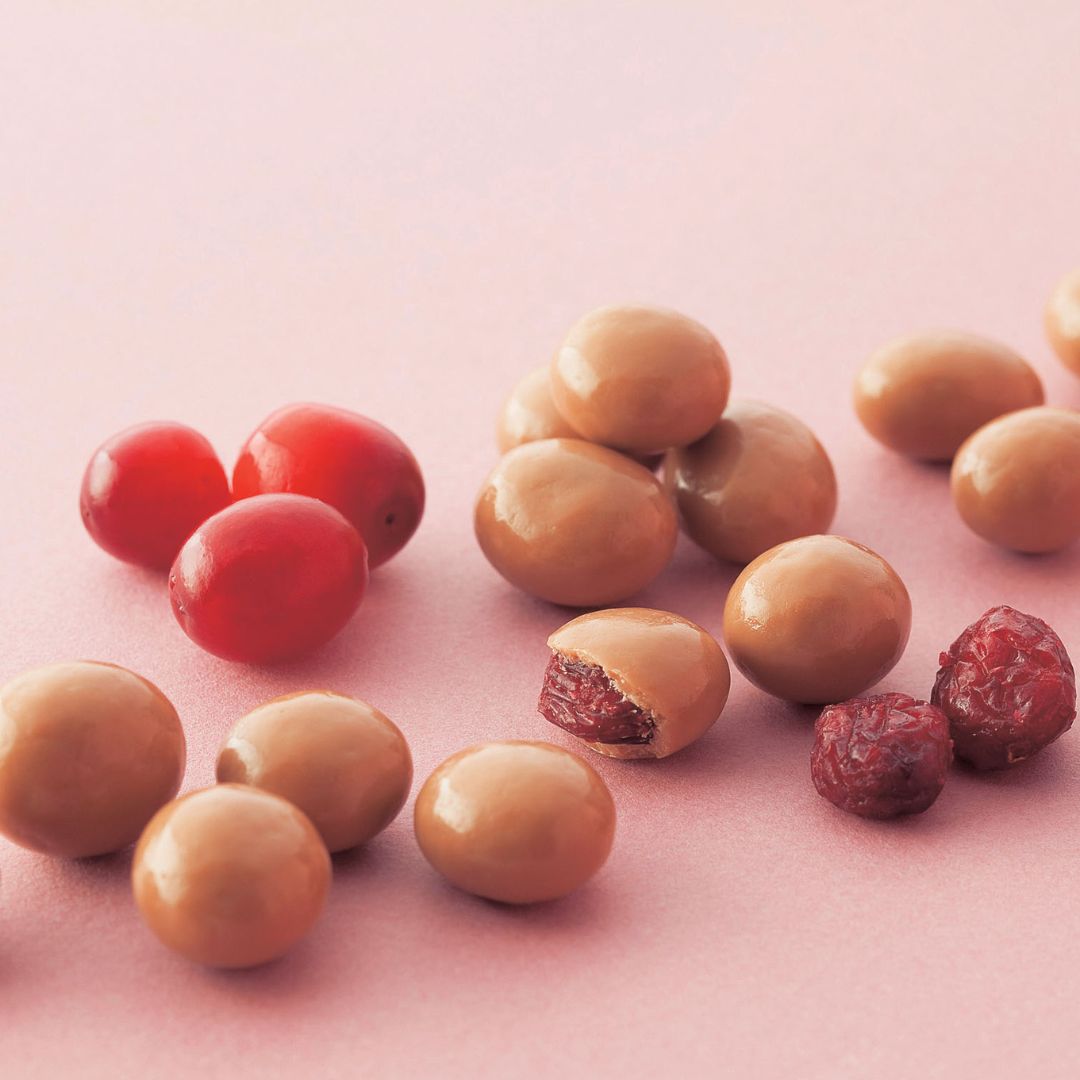 A small pile of dried cranberries next to glossy, milk-chocolate-coated cranberries, one halved to reveal the fruit inside.