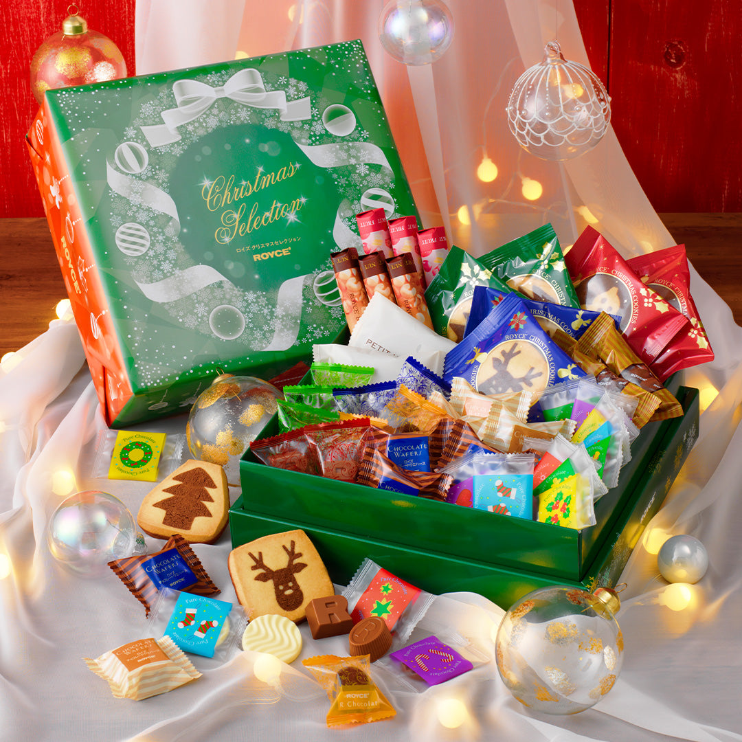 Image shows green printed boxes with illustrations of snowflakes, balls, and ribbons. Accents include wrapped chocolates, clear decor balls, a white cloth, and a red background.