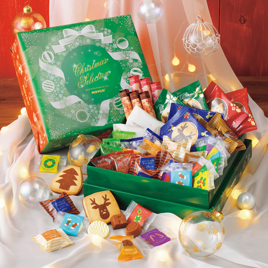 Open Christmas Selection gift box by ROYCE with assorted sweets, including chocolates, cookies, and biscuits, surrounded by festive decorations and warm lights.