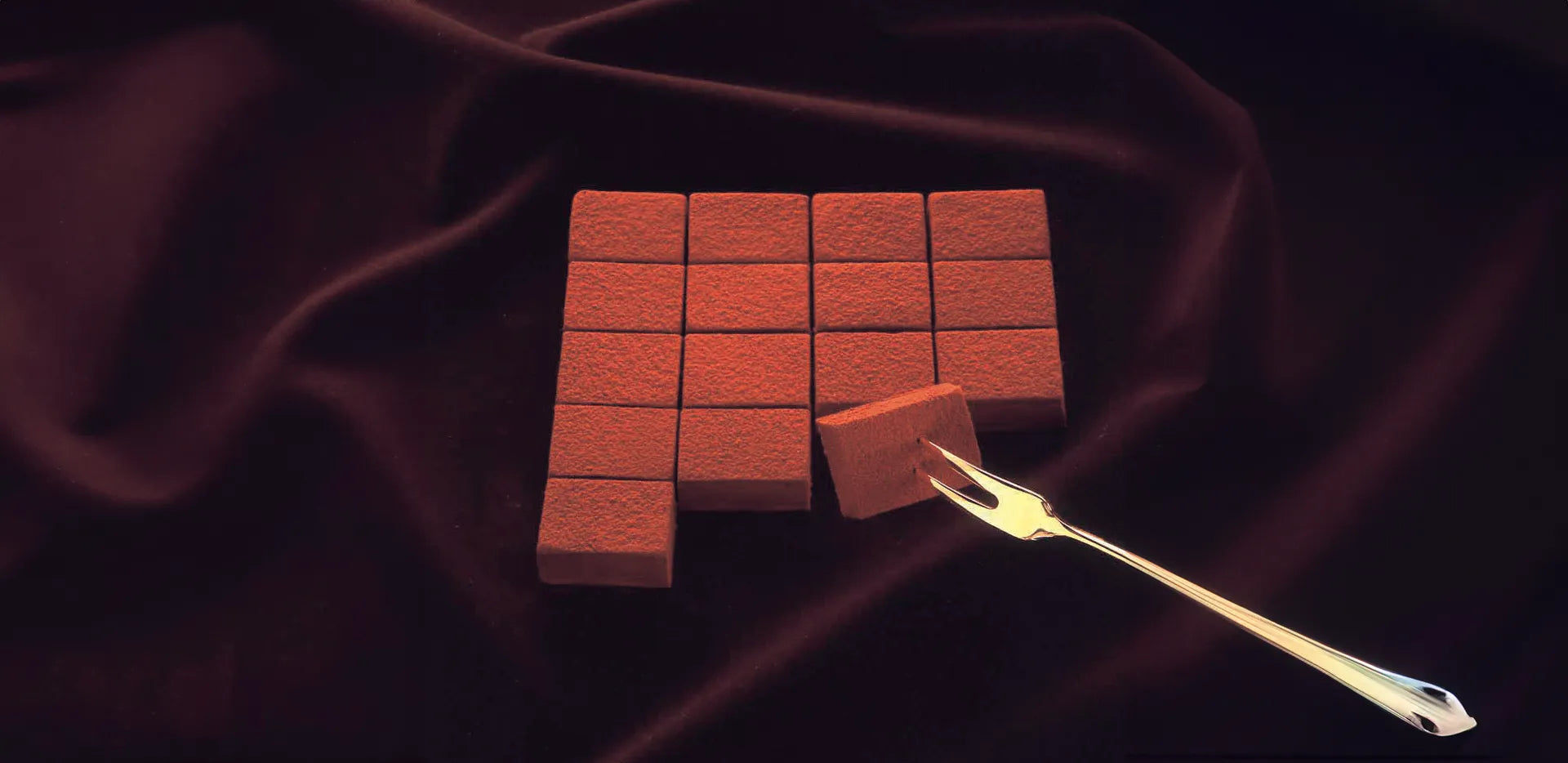 Grid of cocoa-dusted chocolate squares on velvety cloth; a two-pronged fork lifts a piece in the lower right corner.
