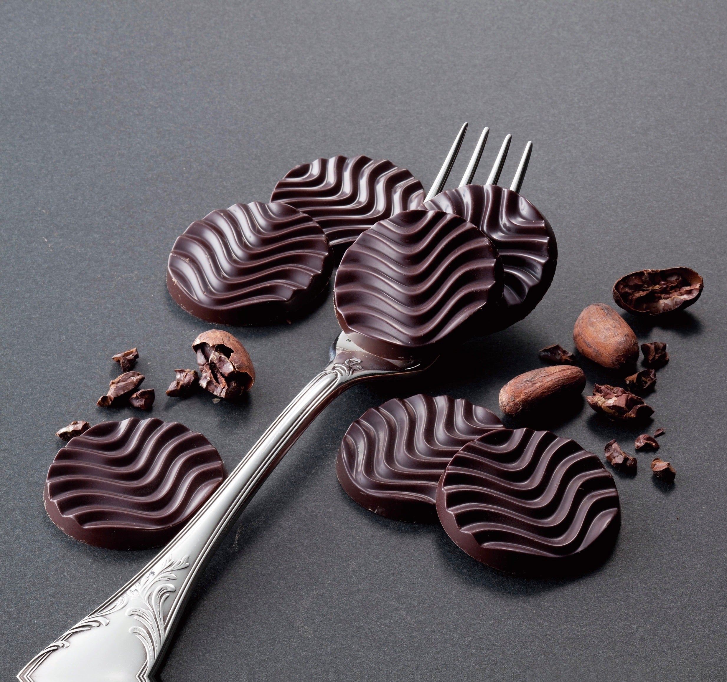 Image shows dark ROYCE' Pure Chocolate discs with a fork and some cacao beans.