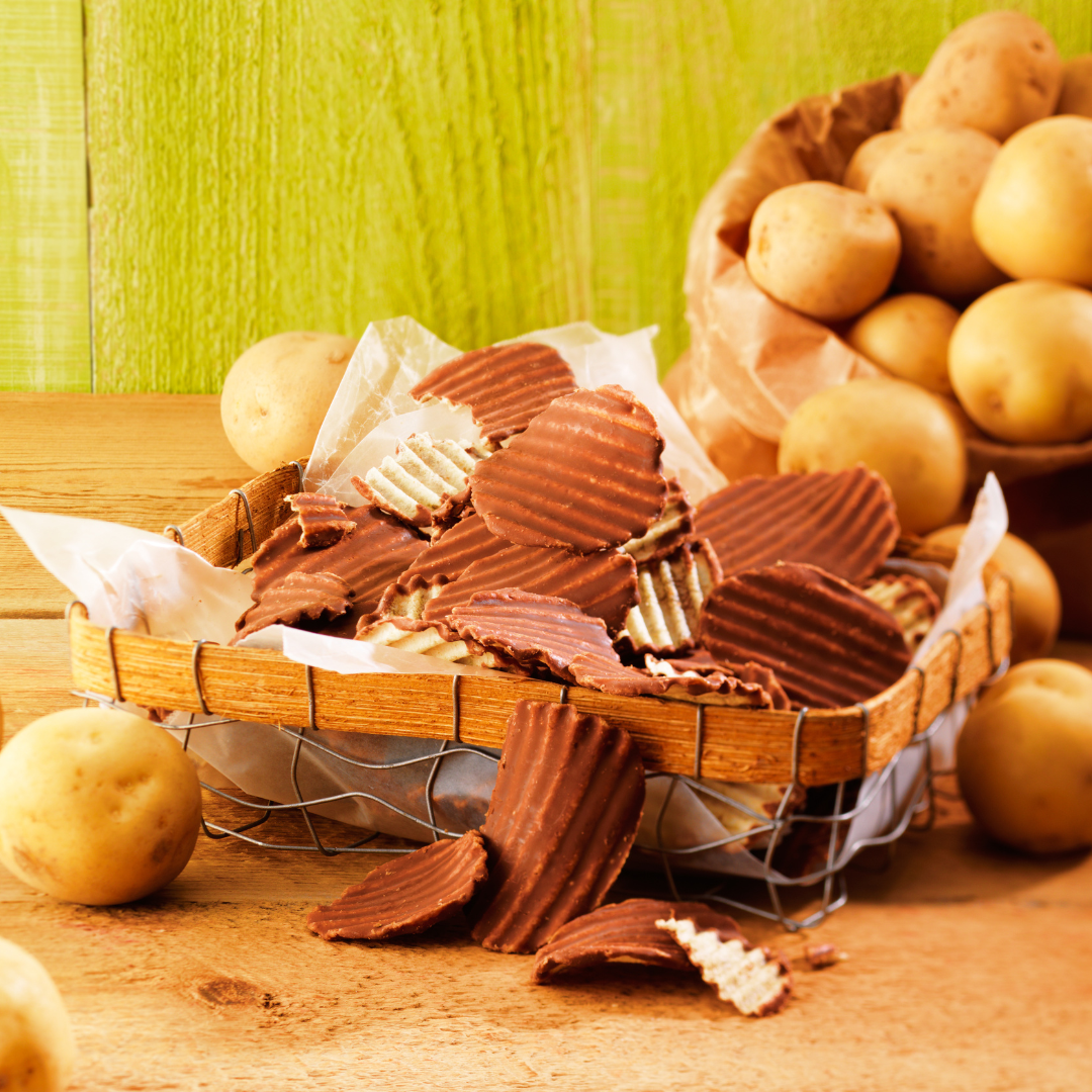 Rustic display of milk chocolate-dipped ridged potato chips in a wire basket, surrounded by whole potatoes on a parchment-lined tabletop.