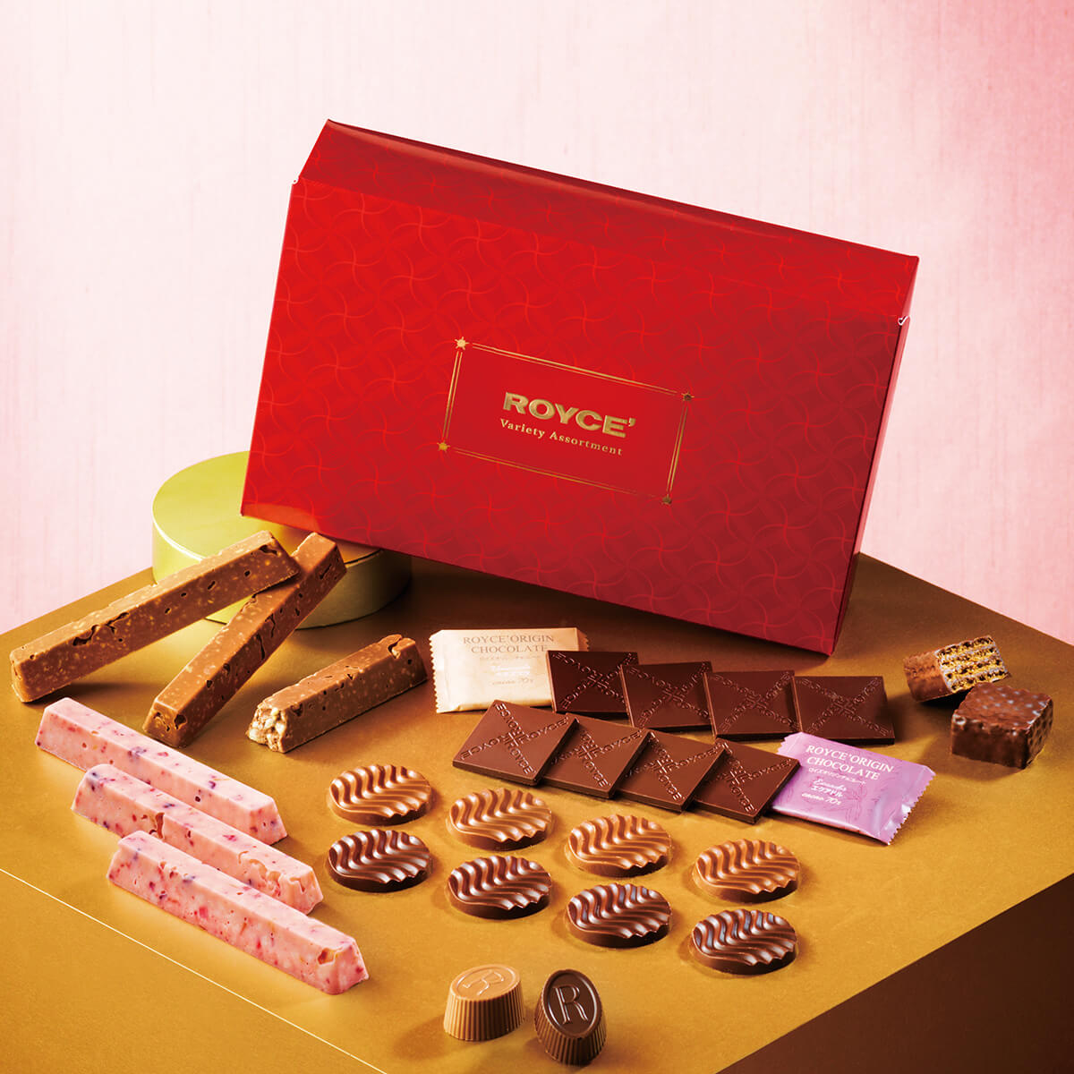 Assorted Royce’ chocolates arranged on a gold surface, featuring bars, squares, discs, pralines, and wafers, centered by a red Royce’ Variety Assortment box.