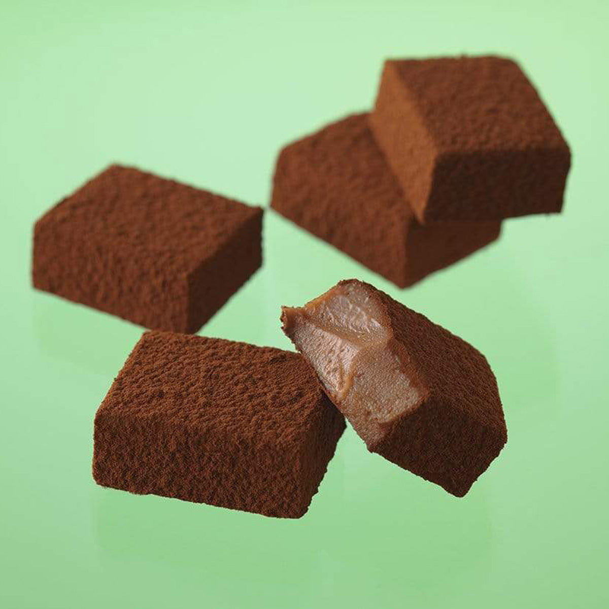 Close-up of Nama chocolate pieces, dusted with cocoa powder; one is cut open, revealing a soft, creamy chocolate interior.