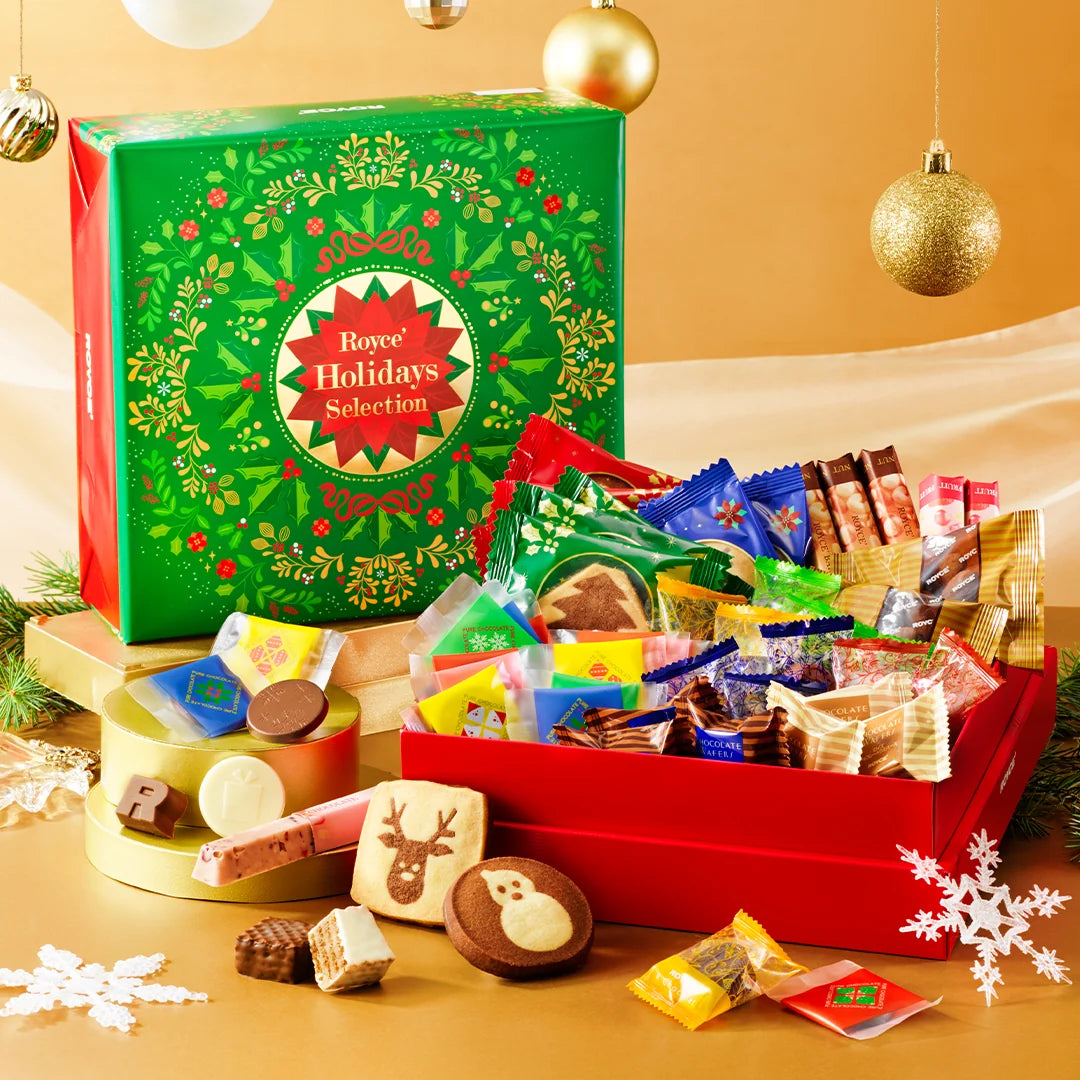 A festive holiday candy and snack selection set displayed with its green branded box and an open red box filled with assorted packaged sweets, cookies, chocolates, and treats on a warm gold background with hanging ornaments.