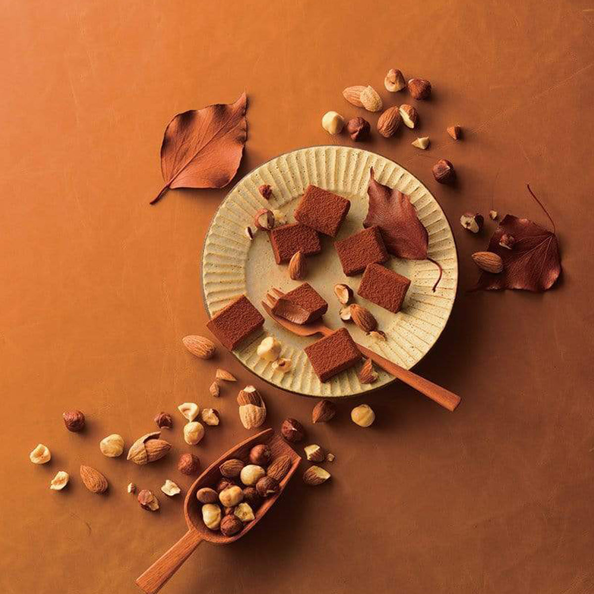 Image shows brown chocolate blocks on a yellow plate. Accents include a brown wooden scoop, a brown wooden fork, loose mixed nuts, and brown leaves. Background surface is in bright brown.