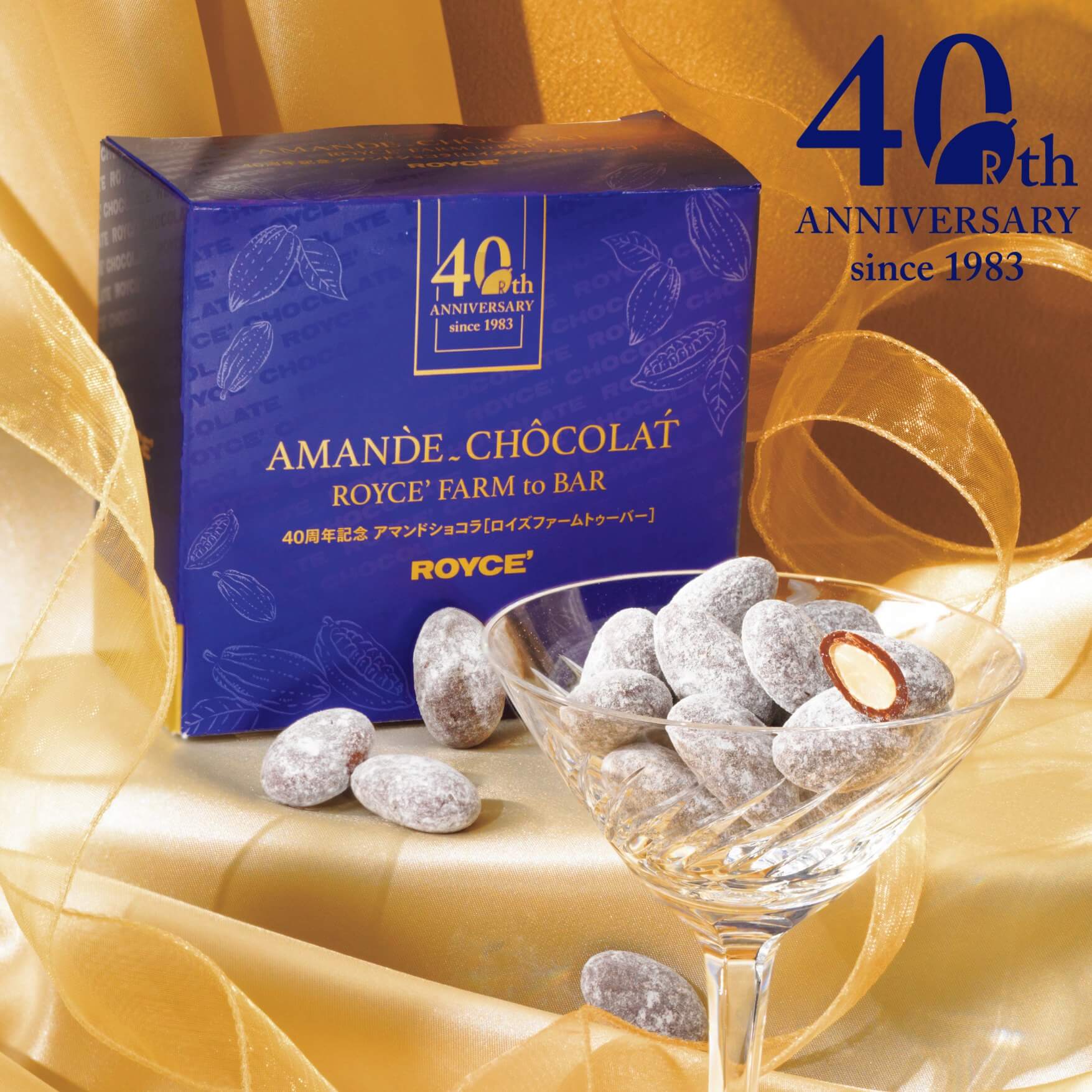 Image shows a cocktail glass with chocolate-coated almonds coated with white sugar powder. Blue box behind has text saying 40th Anniversary since 1983 Amande Chocolat ROYCE' Farm to Bar. ROYCE'. Blue text on upper right says 40th Anniversary since 1983. Background is in gold.