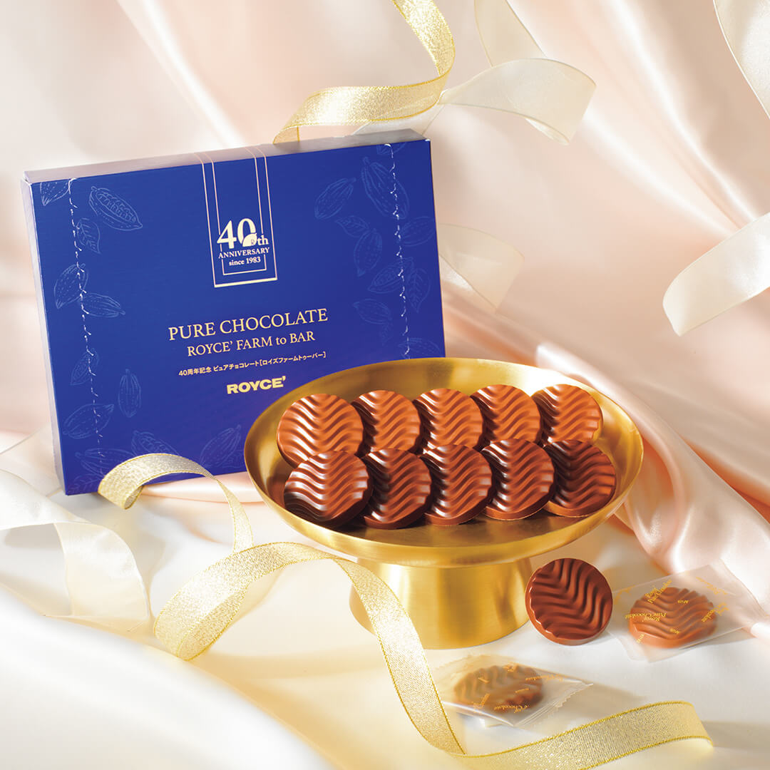 Image shows a blue box with a gold plate and brown chocolate discs. Text says 40th Anniversary since 1983. 40th Pure Chocolate ROYCE' Farm to Bar. ROYCE'.