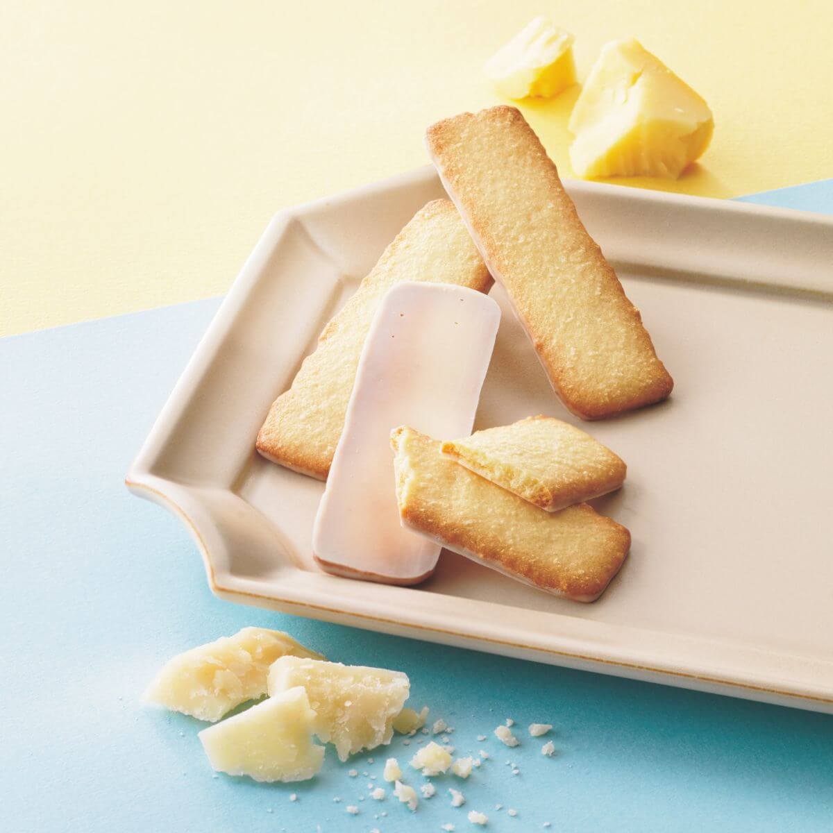 Baton Cookies Fromage (25 Pcs) displayed on a plate with cheese crumbs and butter cookies, featuring coconut, Parmesan, and Gouda flavors, coated in white chocolate.