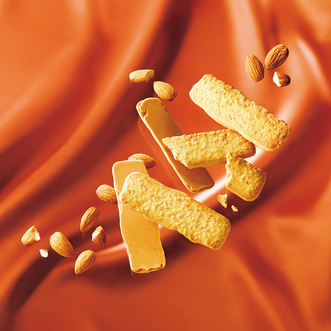 Baton Cookies "Almond" - Golden-brown almond cookies and whole almonds floating against a draped orange fabric background.