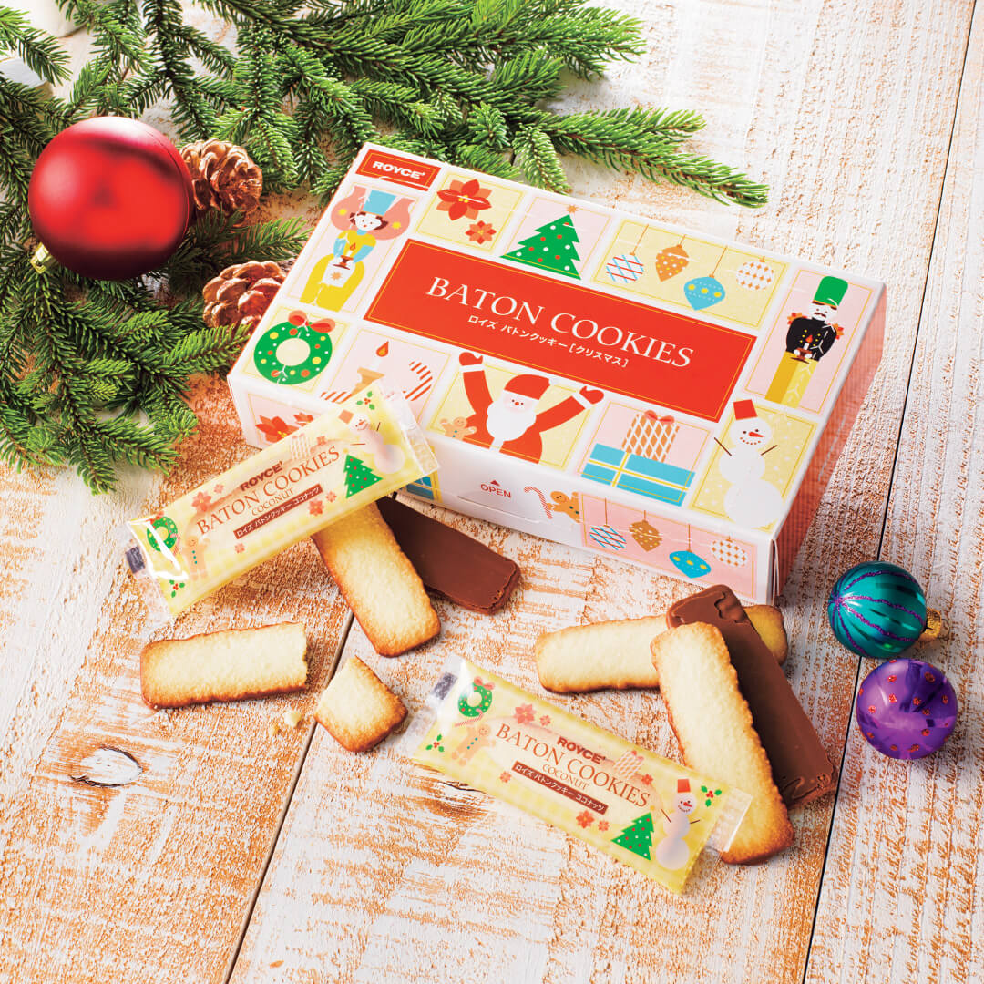 Festive display of ROYCE Baton Cookies Christmas: colorful box with holiday motifs, surrounded by butter biscuits and wrapped packets, framed by evergreen sprigs and pine cones.