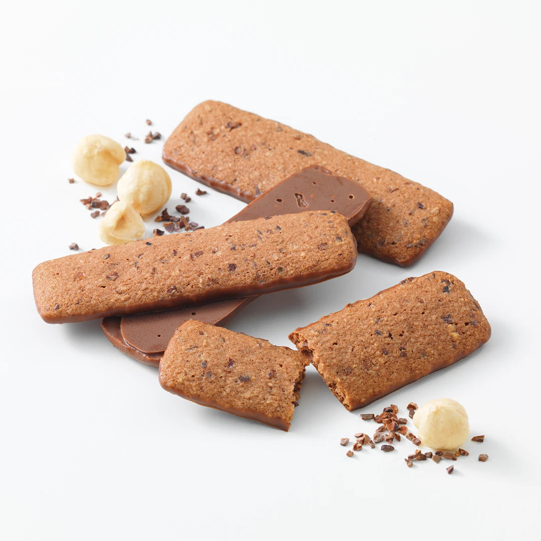 Baton Cookies Hazel Cacao (25 Pcs)—rectangular cookie bars half-dipped in milk chocolate, surrounded by whole hazelnuts and cocoa nibs on a white background.