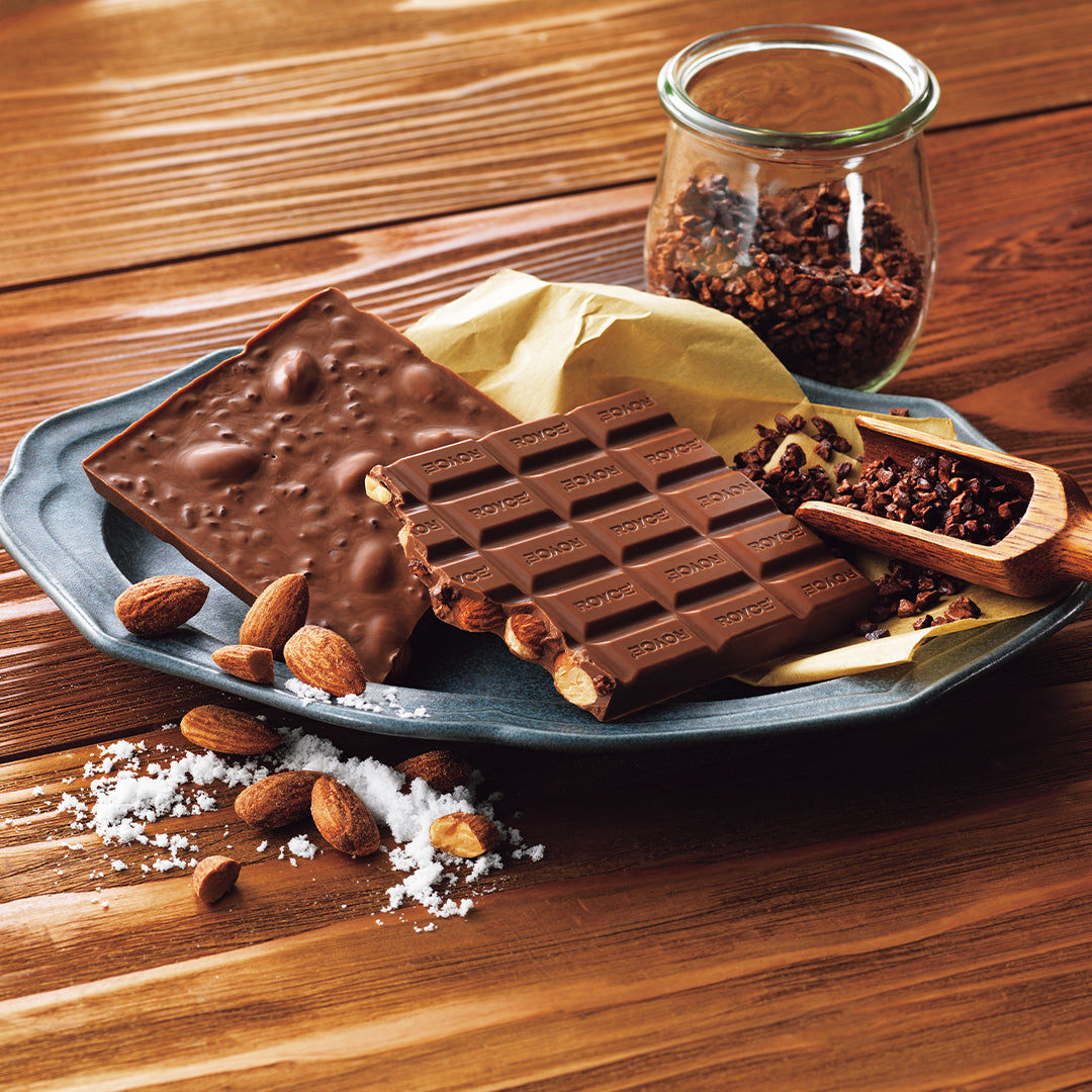 A styled food product shot showing chocolate bars with almonds and sea salt on a blue-gray plate, with a small glass jar of cacao beans and a wooden scoop on a wooden tabletop.
