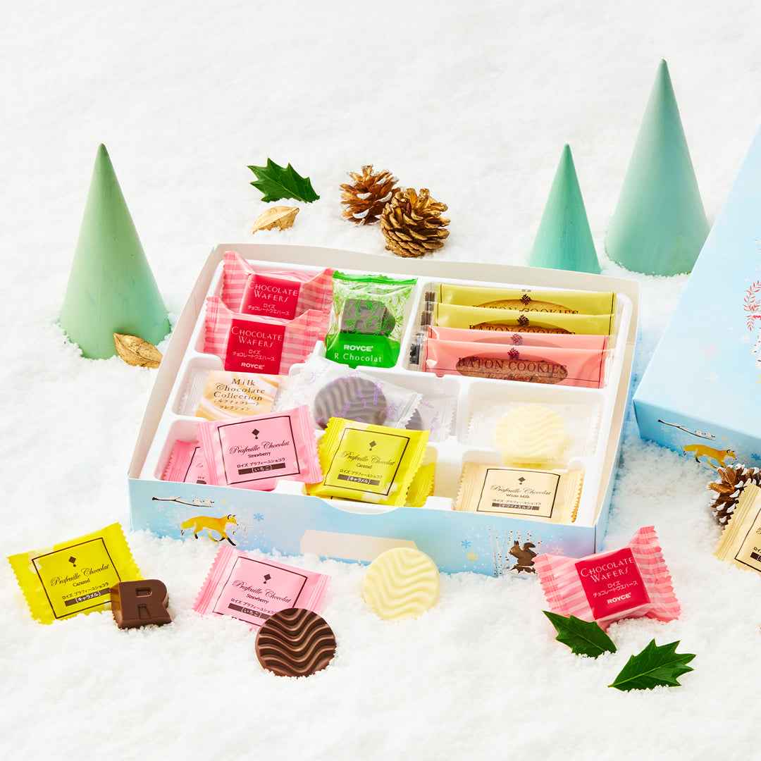 An assortment of packaged chocolates and sweets displayed in an open gift box on a snowy surface, surrounded by miniature green trees, pinecones, and holly leaves. The sweets come in pink, yellow, green, and white wrappers, with some unwrapped chocolates showing brown and cream filling.