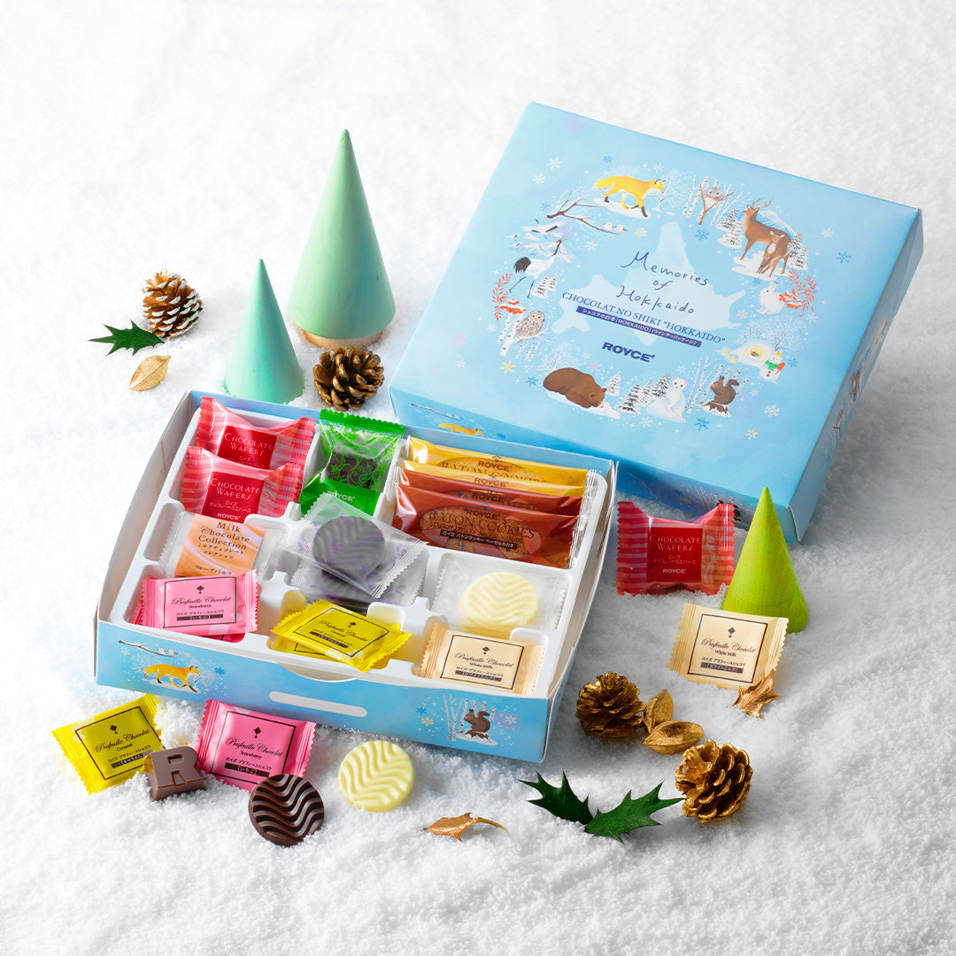 Image shows a blue box with illustrations of a fox, deer, a crane, owls, a bear, squirrels, a snowman, a cat, a rabbit, and snow-covered plants. Text says Memories of Hokkaido. Chocolat No Shiki "Hokkaido". ROYCE'. Lower box has confections in different shapes and colors. Other text are the words Baton Cookies, Chocolate Wafers, Milk Chocolate Collection, ROYCE', and Prafeuille Chocolat. Setting is a white snow-covered ground with accents of miniature green trees, brown acorns, and loose chocolates.