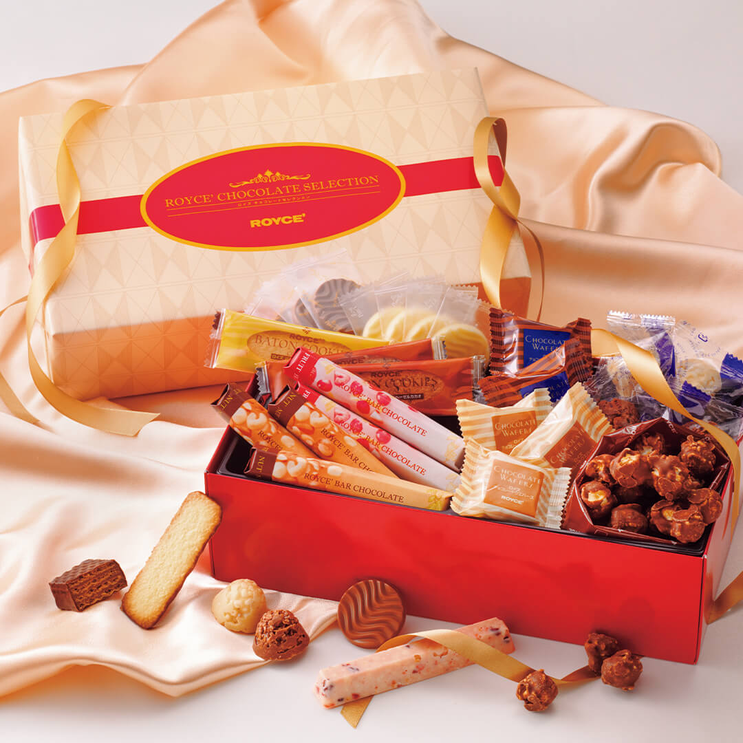 ROYCE' Chocolate - Chocolate Selection "ROYCE' Sweet Season" - Image shows two boxes on beige silk cloth. Top left box is in nude and pink colors with text saying ROYCE' Chocolate Selection ROYCE'. Below right shows red box filled with individually-wrapped confections and open chocolates in varying colors. Accents include red ribbons gold ribbons and loose confections.