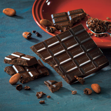 ROYCE' Chocolate - Chocolate Bar "Cacao Nibs" - Image shows a dark chocolate bar with the words ROYCE' etched into it. Accents include loose and crushed cacao beans, a red plate, and a metal spoon. Background is in blue.