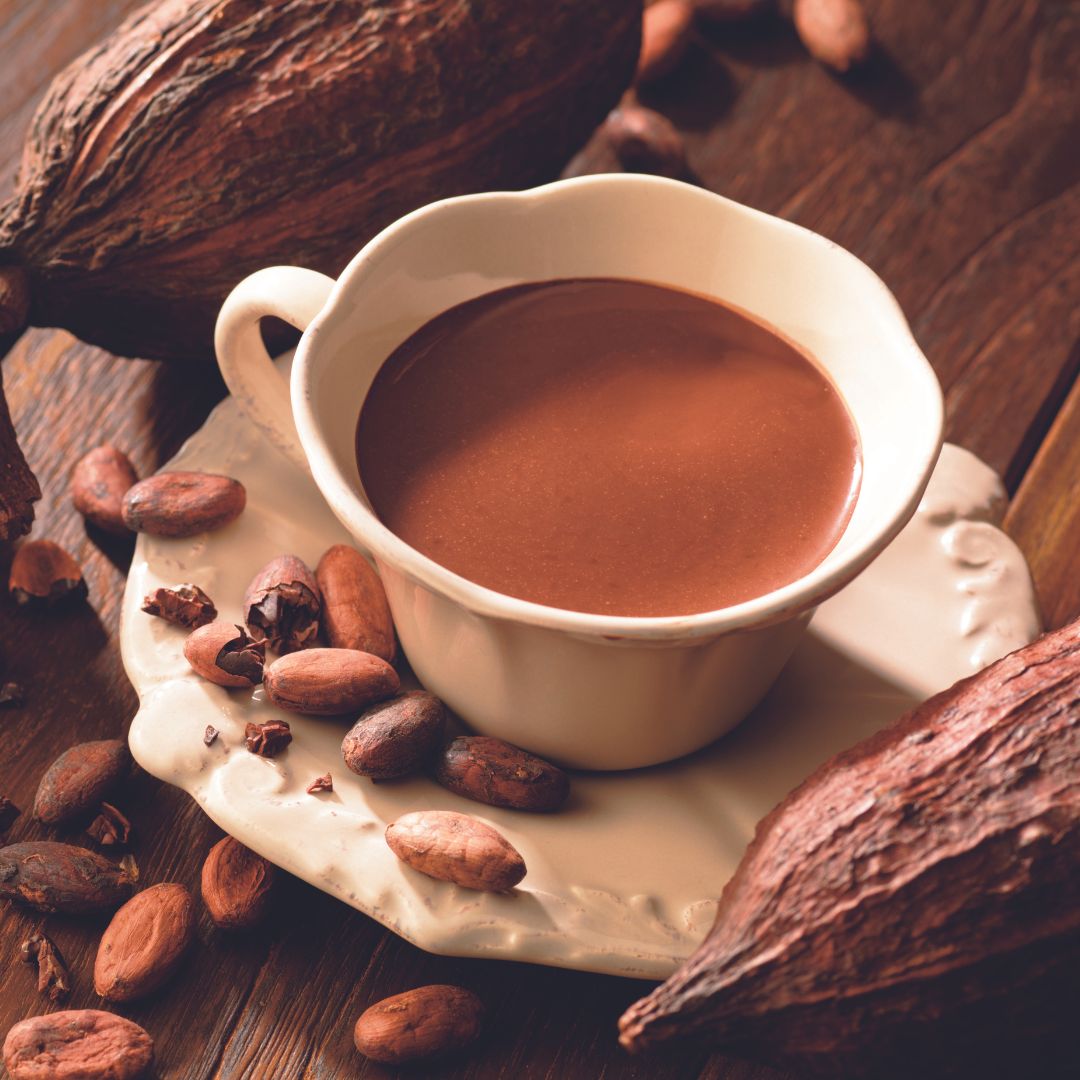 Image shows a cup filled with hot chocolate on a plate. Accents include cacao fruits and loose cacao beans.