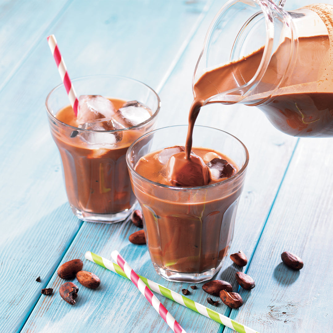 Image shows glasses with ice chocolate on a table with straws and cacao beans.