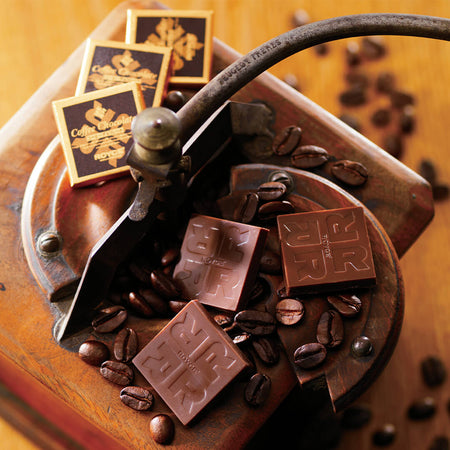 ROYCE' Chocolate - Coffee Chocolate - Image shows brown chocolate squares embossed with the letter R and word ROYCE'. Also included in the picture are loose coffee beans on a wooden platform and individually-wrapped chocolates. 