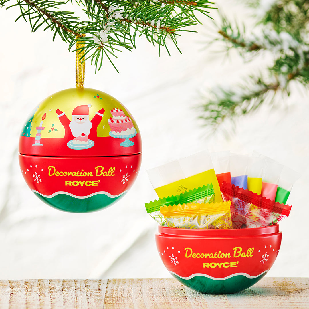 A festive red and green Christmas decoration ball ornament hanging from a pine branch, shown beside a matching bowl filled with colorful wrapped candies. The ball and bowl are branded ROYCE.
