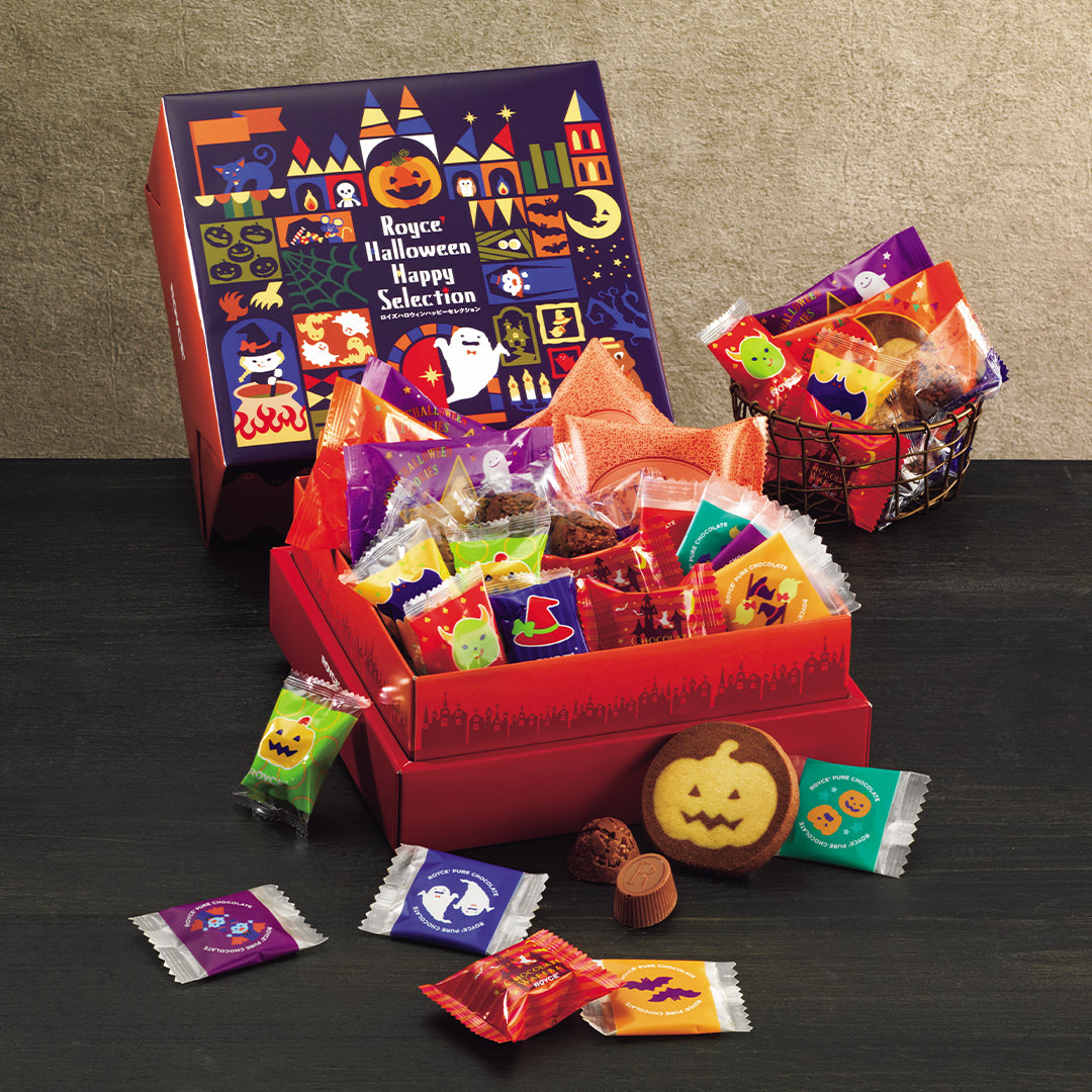 ROYCE' Halloween Happy Selection - A Halloween-themed gift set from ROYCE' featuring a large illustrated box, an open red box filled with assorted packaged confections, and several loose chocolates scattered on a dark tabletop against a beige wall.