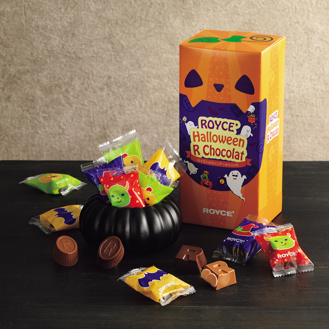 ROYCE' Halloween R Chocolat - Image shows chocolate assortments with a large orange pumpkin-shaped box, a small black pumpkin container filled with individually wrapped chocolates, and several loose chocolates arranged on a dark tabletop against a textured beige wall.