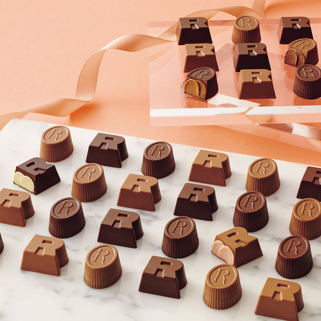 ROYCE' Halloween R Chocolat - Image shows an assortment of milk and dark chocolate candies shaped like the letter 'R', arranged on a white marble surface with a peach background and a matching gift box in the upper right.