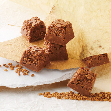 ROYCE' Kyoto Kin Goma Crunch Chocolate - Image shows brown chocolate cubes on a surface of white and gold paper. Accents include loose sesames.
