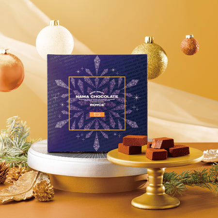 Image shows a blue printed box with a snowflake illustration. Accents include gold and white plates, brown chocolate blocks, leaves, and decorative orbs and leaves in the colors of gold and bronze. Background is in various hues of gold color.