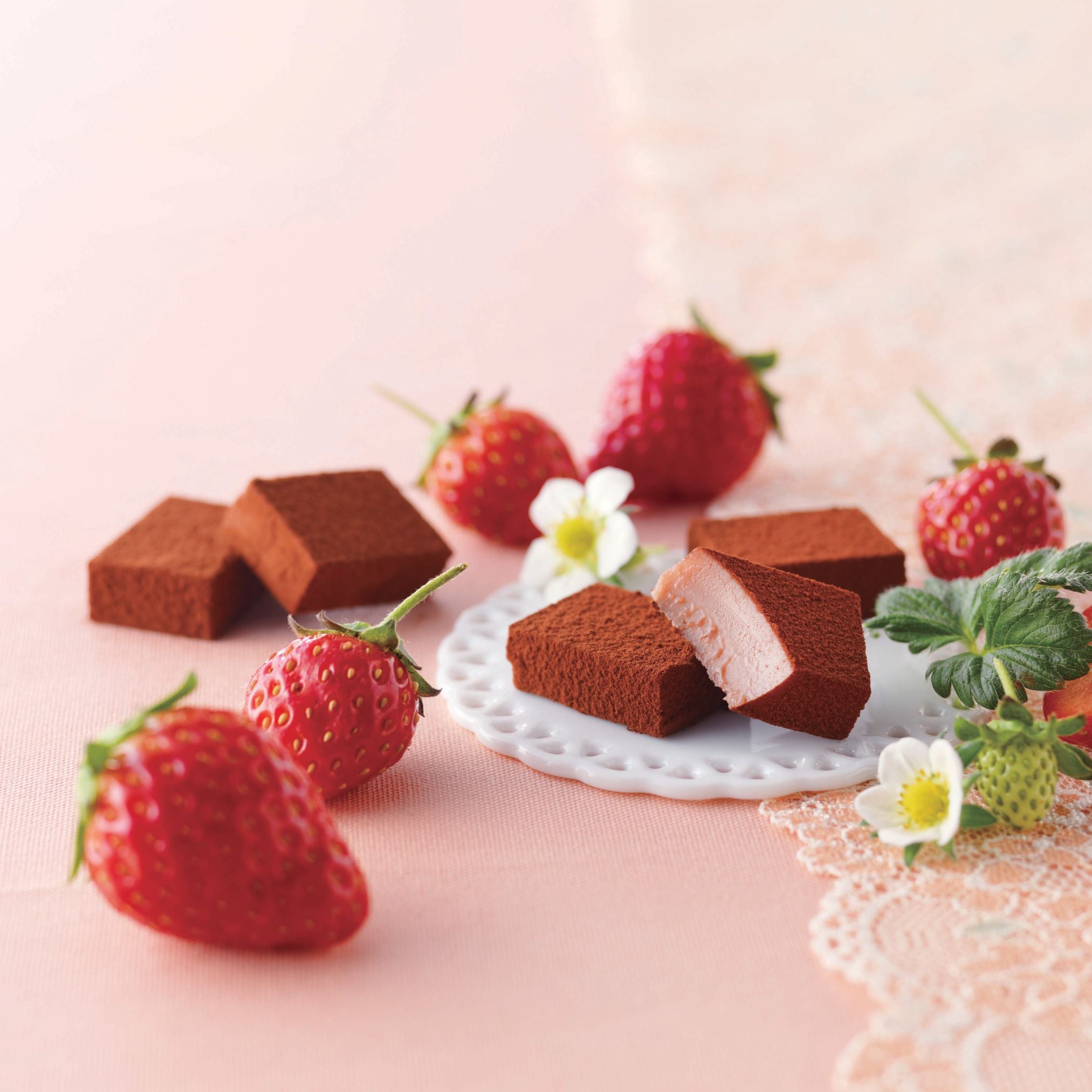 ROYCE' Chocolate - Nama Chocolate "Strawberry" - Image shows brown blocks of chocolates with accents of a white coaster, red strawberries, and green leaves. Background is in pink.