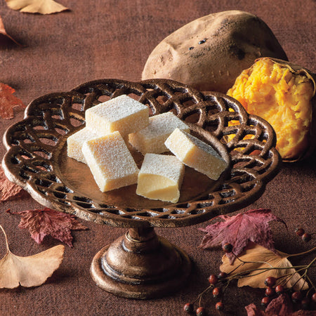 ROYCE' Chocolate - Nama Chocolate "Roasted Sweet Potato" - Image shows white chocolate blocks in a metallic plate on a brown surface. Accents include loose leaves and sweet potatoes.