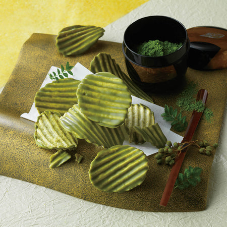 ROYCE' Kyoto Potatochip Chocolate "Uji Matcha & Sansho" - Image shows potato chips coated with green chocolate on a plate. Accents include loose grape peppers, a wooden stick, some leaves, and a black tea cup with green matcha powder inside.