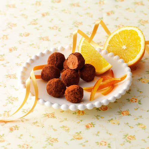 Petite Truffe Orange chocolates on a scalloped plate with cocoa dust, orange wedges, and ribbon; one truffle cut to show creamy orange-infused ganache.