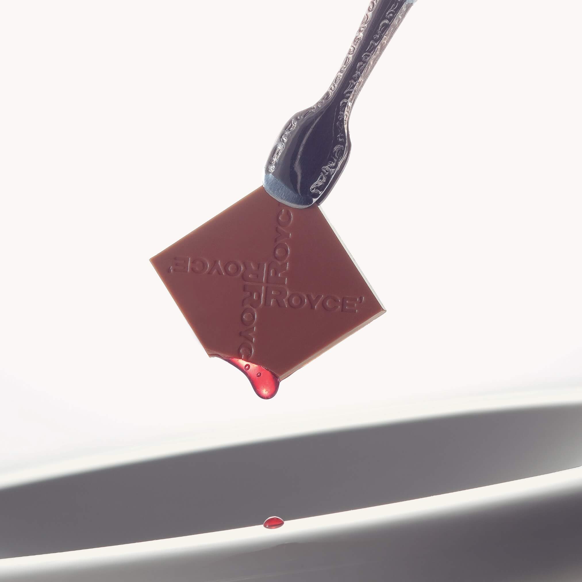 ROYCE' Chocolate - Prafeuille Chocolat "Berry Cube" - Image shows a silver tong holding a brown chocolate square with red sauce and engraved with the words "ROYCE'". Accents include a white plate-like surface. Background is in color white.