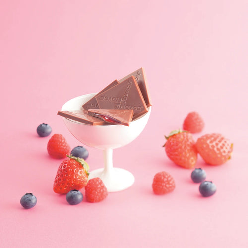 Prafeuille Chocolat Berry Cube: Triangular chocolate pieces with red-berry filling in a glass, surrounded by strawberries, raspberries, and blueberries on a pink surface.