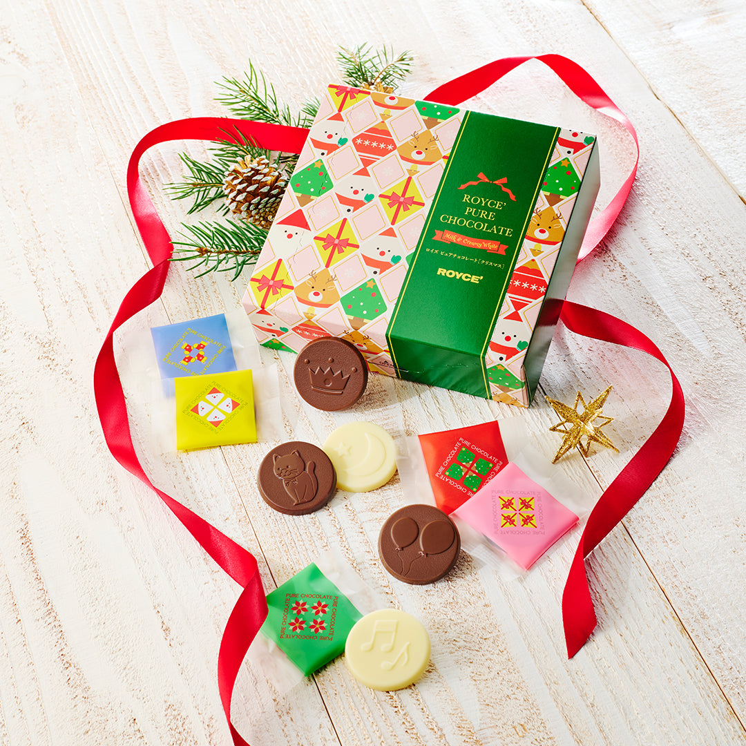 A boxed assortment of chocolate discs arranged on a light wood surface with a red ribbon, evergreen branches, a pinecone, and gold star decorations.
