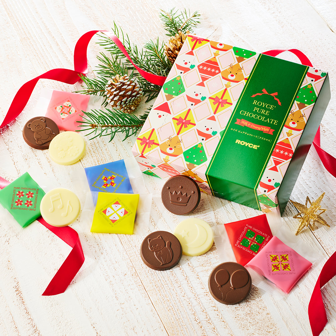 A boxed assortment of chocolate discs arranged on a light wood surface with a red ribbon, evergreen branches, a pinecone, and gold star decorations.