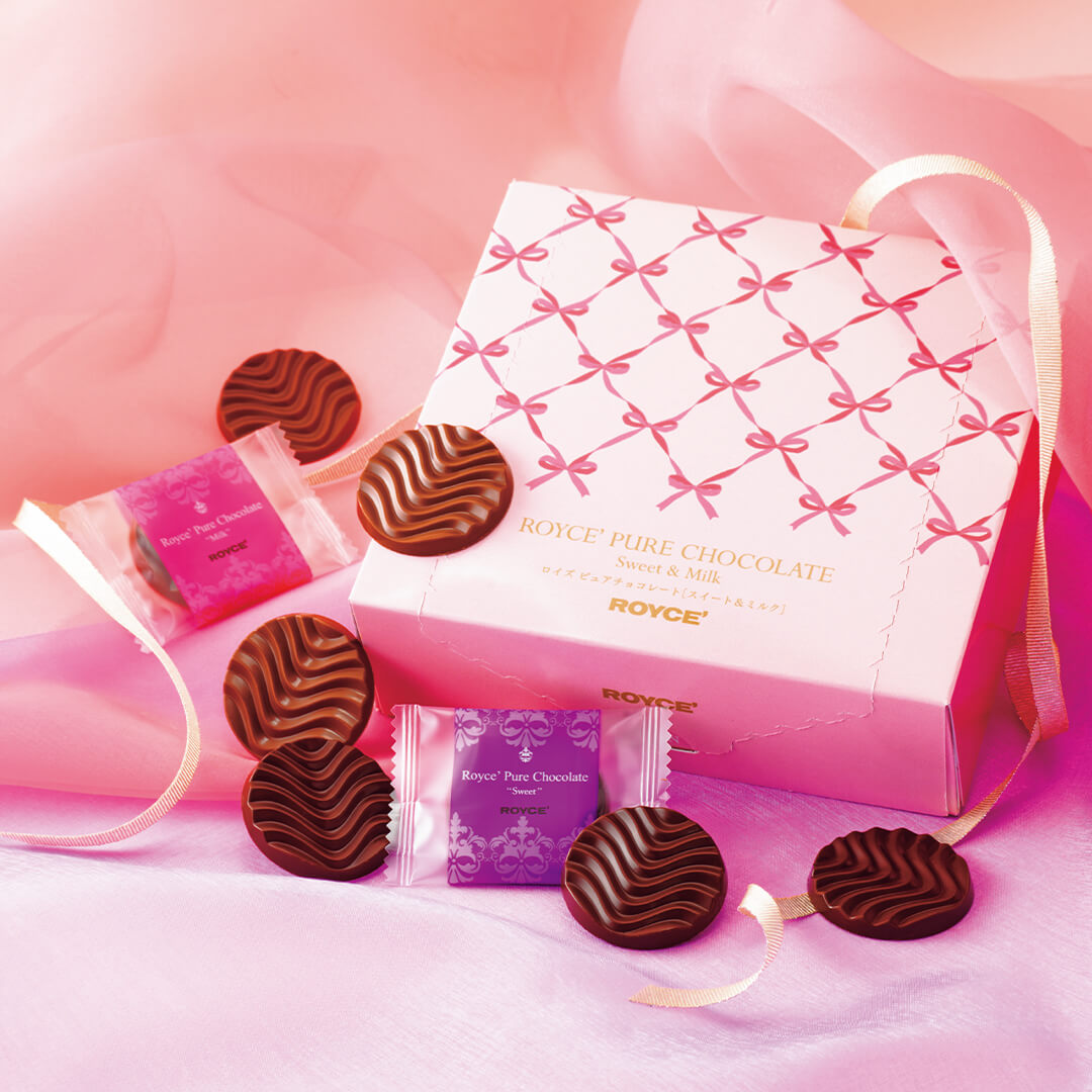 ROYCE' Chocolate - Pure Chocolate "Sweet & Milk" (Gift) - Image shows a pink box with ribbon prints and gold text saying ROYCE' Pure Chocolate Sweet & Milk ROYCE'. Accents include a gold ribbon, individually-wrapped chocolates in violet and pink, as well as brown chocolate discs with a waved texture. Background features a pink waved cloth on top and a silky violet cloth below.