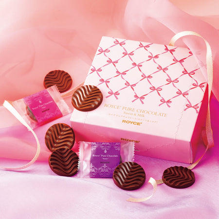 ROYCE' Chocolate - Pure Chocolate "Sweet & Milk" (Gift) - Image shows a pink box with ribbon prints and gold text saying ROYCE' Pure Chocolate Sweet & Milk ROYCE'. Accents include a gold ribbon, individually-wrapped chocolates in violet and pink, as well as brown chocolate discs with a waved texture. Background features a pink waved cloth on top and a silky violet cloth below.
