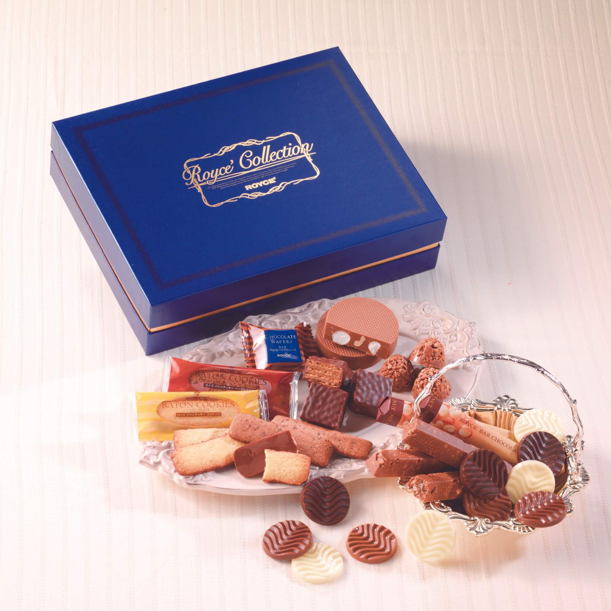 ROYCE' Chocolate - ROYCE' Collection "Blue" - Image shows on top left a blue box. Gold text says ROYCE' Collection ROYCE'. Below the box are a mix of confections in various shapes and colors, as arranged in accents such as a glass plate and silver bowl. Background and surface is a white tablecloth.