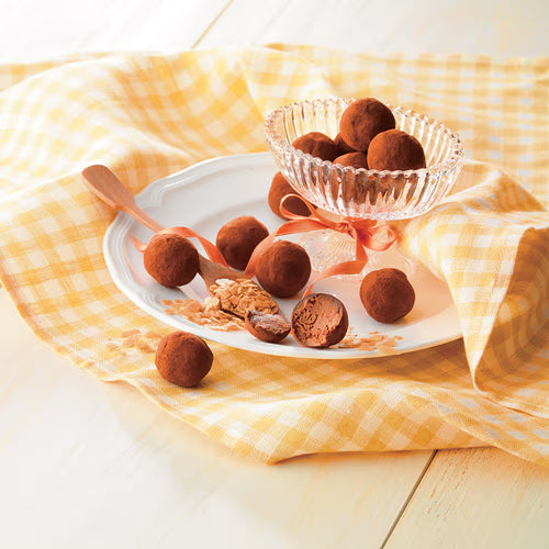 ROYCE’ Feuillantine Truffe “Gianduja” on a white plate, showcasing cocoa-dusted truffles with one halved, revealing a fudgy interior, beside a wooden spoon on gingham cloth.