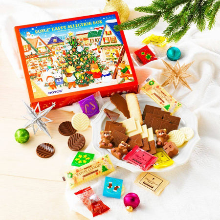 ROYCE' Chocolate - ROYCE' Happy Selection Box - Image shows a printed box on top left with illustrations of animals, houses, and a decorated tree. Text says ROYCE' Happy Selection Box. Accents include chocolates and confections, a white plate, decorative orbs and stars, a white tablecloth, and green mistletoe.