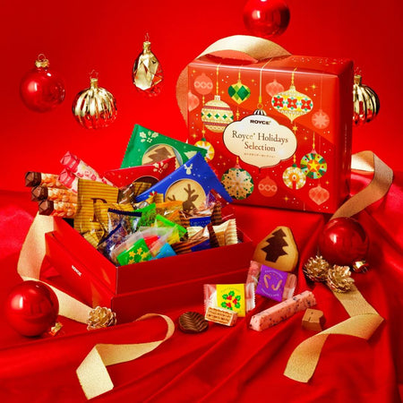ROYCE' Chocolate - ROYCE' Holidays Selection (32 Pcs) - Image shows a printed red box with illustrations of decoration balls and lamps. Text says ROYCE' Holidays Selection. Also seen is an open red box with varying individually-wrapped confections. Accents include a decorative orbs, gold acorns, gold ribbon, and loose confections. Background is in red.